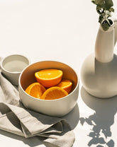A white stoneware serving bowl filled with orange slices, placed on a table with a grey napkin and a white vase in the background.